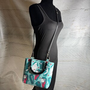 K&ouml;nnte beinhalten: Eine t&uuml;rkisgr&uuml;ne Handtasche mit schwarzem Griff und Riemen, mit einem Muster aus Zeichentrickfiguren, S&auml;rgen und roten Punkten. Die rechteckige Tasche hat eine strukturierte Form und silberfarbene Beschl&auml;ge.