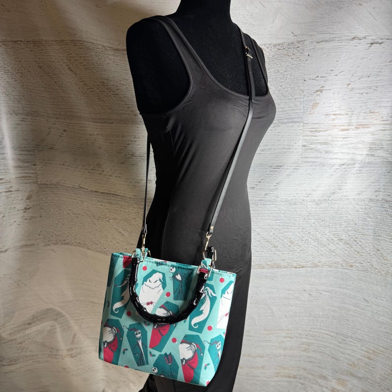 K&ouml;nnte beinhalten: Eine t&uuml;rkisgr&uuml;ne Handtasche mit schwarzem Griff und Riemen, mit einem Muster aus Zeichentrickfiguren, S&auml;rgen und roten Punkten. Die rechteckige Tasche hat eine strukturierte Form und silberfarbene Beschl&auml;ge.