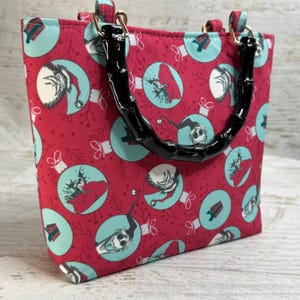 Puede incluir: Un bolso tote rojo con un asa de bamb&uacute; negro. El bolso presenta un patr&oacute;n repetido de adornos navide&ntilde;os de color turquesa con personajes de dibujos animados. El asa est&aacute; sujeta con herrajes dorados.