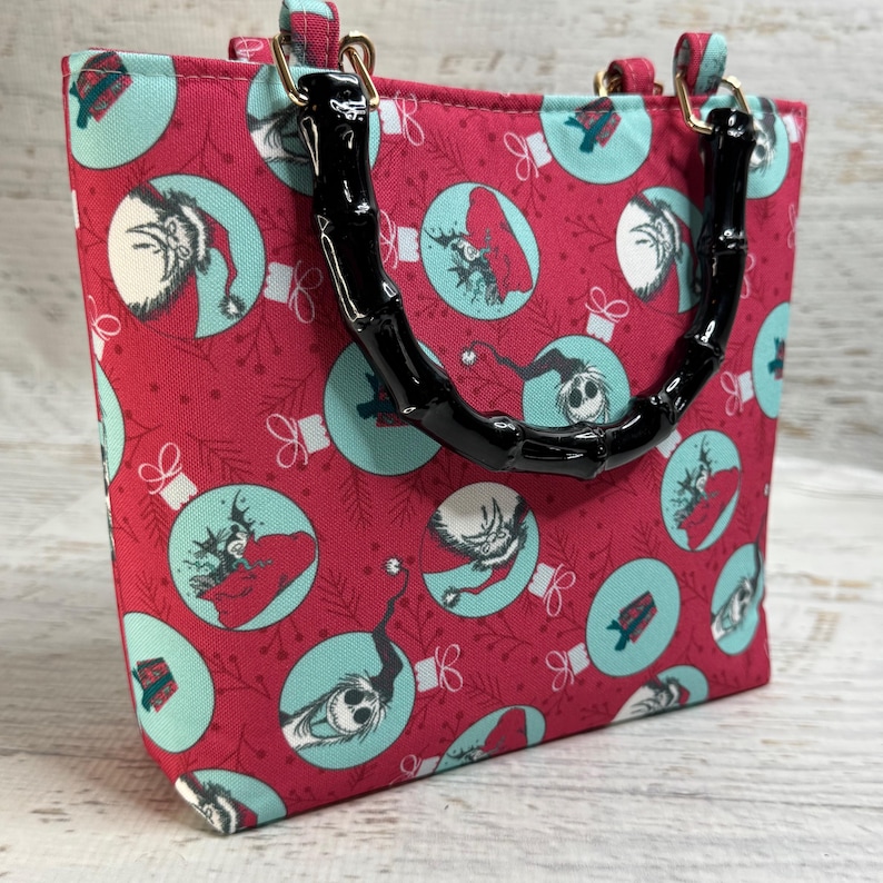 Puede incluir: Un bolso tote rojo con un asa de bamb&uacute; negro. El bolso presenta un patr&oacute;n repetido de adornos navide&ntilde;os de color turquesa con personajes de dibujos animados. El asa est&aacute; sujeta con herrajes dorados.