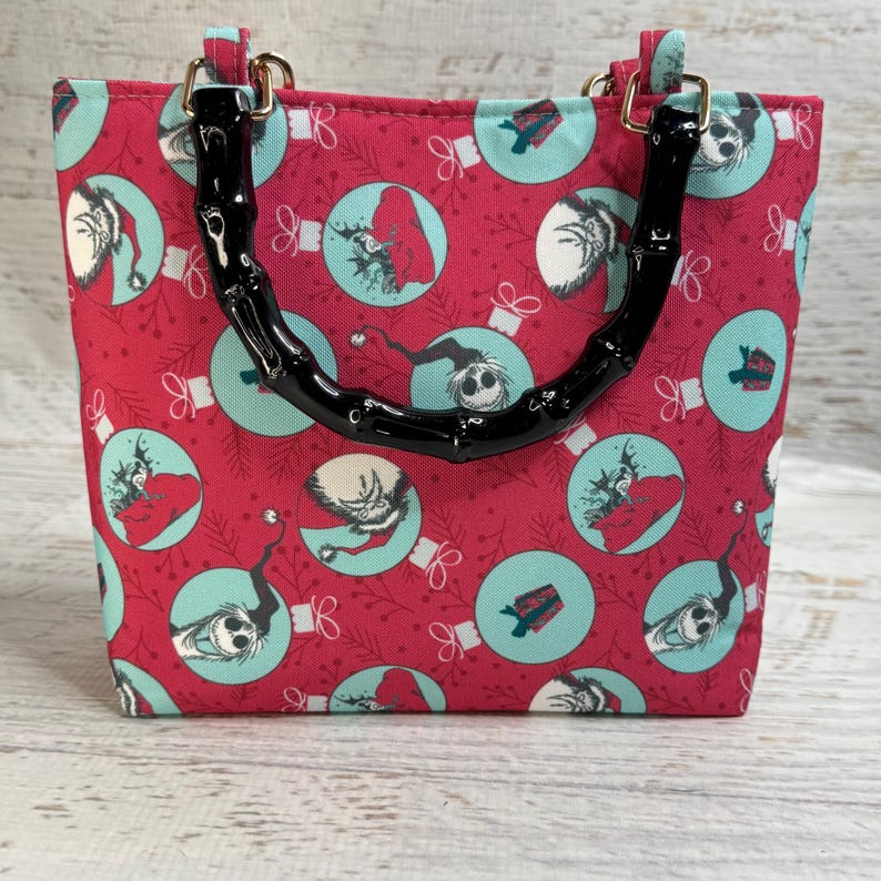 Puede incluir: Un bolso tote rojo con un patr&oacute;n repetido de adornos navide&ntilde;os de color turquesa con personajes. El bolso tiene asas negras estilo bamb&uacute; y herrajes dorados. El bolso tiene forma cuadrada.