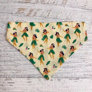 May include: A yellow bandana with a pattern of hula dancers in green skirts and floral leis. The background is a light yellow with green leaves and pink flowers.