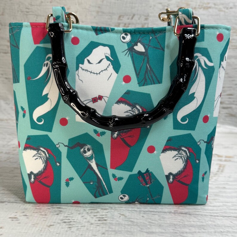 K&ouml;nnte beinhalten: Eine kleine Tragetasche mit t&uuml;rkisblauem Hintergrund mit Charakteren aus The Nightmare Before Christmas. Die Tasche hat einen schwarzen Bambusgriff und goldfarbene Beschl&auml;ge. Das Design umfasst Bilder von Jack Skellington, Oogie Boogie und Santa Jack.