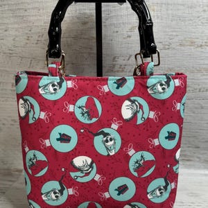 Puede incluir: Un peque&ntilde;o bolso tote con un fondo rojo y un patr&oacute;n repetido de adornos de color turquesa con personajes. El bolso tiene un asa negra estilo bamb&uacute; y herrajes dorados. El bolso est&aacute; sobre un soporte negro.