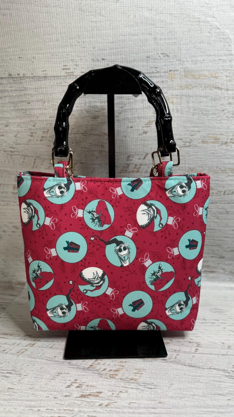 Puede incluir: Un peque&ntilde;o bolso tote con un fondo rojo y un patr&oacute;n repetido de adornos de color turquesa con personajes. El bolso tiene un asa negra estilo bamb&uacute; y herrajes dorados. El bolso est&aacute; sobre un soporte negro.