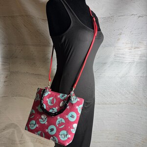 Puede incluir: Bolso tote con estampado rojo y turquesa con asa negra y correa para el hombro roja. El bolso presenta un patr&oacute;n repetido de adornos de calaveras sobre un fondo rojo. El bolso se muestra en un maniqu&iacute; negro.