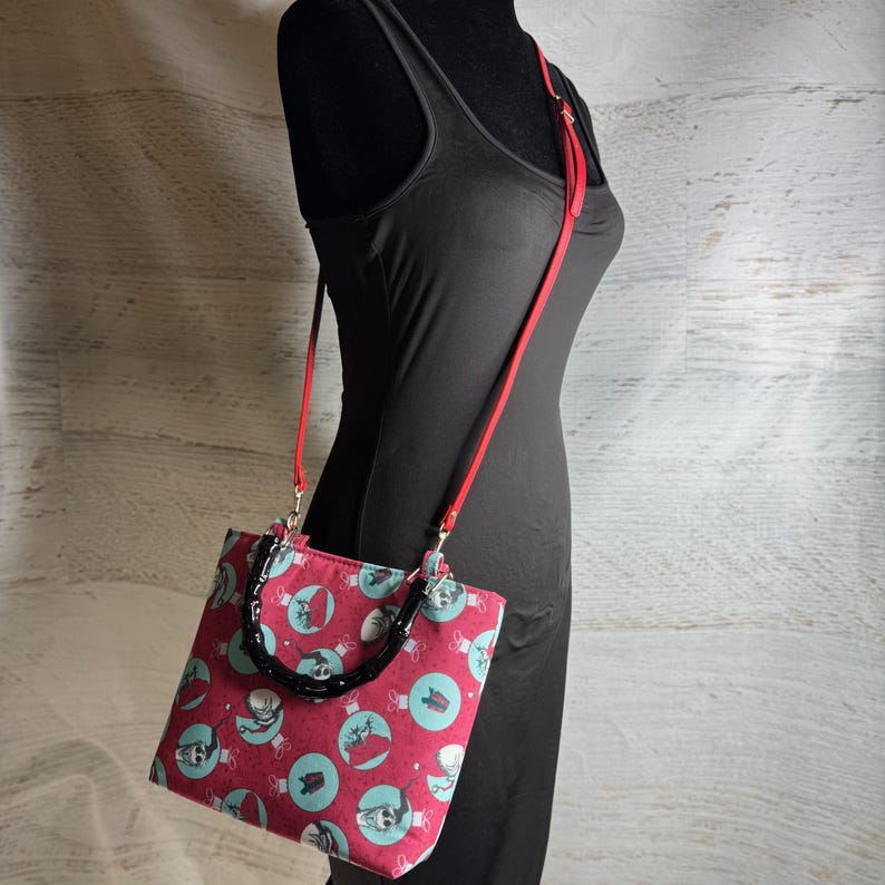 Puede incluir: Bolso tote con estampado rojo y turquesa con asa negra y correa para el hombro roja. El bolso presenta un patr&oacute;n repetido de adornos de calaveras sobre un fondo rojo. El bolso se muestra en un maniqu&iacute; negro.