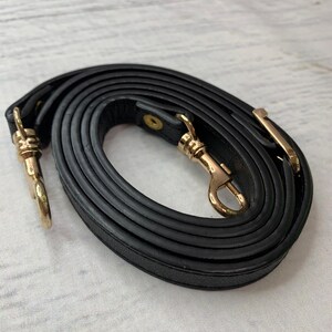 K&ouml;nnte beinhalten: Ein schwarzer Leder-Handtaschenriemen mit goldfarbenen Metallverschl&uuml;ssen und Beschl&auml;gen. Der Riemen ist aufgerollt und zeigt seine L&auml;nge und die Details der Verschl&uuml;sse.