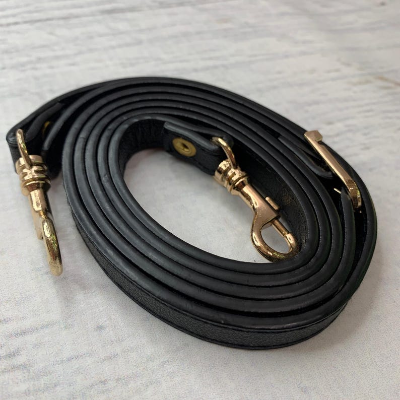 K&ouml;nnte beinhalten: Ein schwarzer Leder-Handtaschenriemen mit goldfarbenen Metallverschl&uuml;ssen und Beschl&auml;gen. Der Riemen ist aufgerollt und zeigt seine L&auml;nge und die Details der Verschl&uuml;sse.