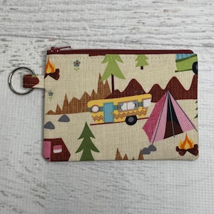 May include: A beige camping-themed coin purse with a zipper closure and a key ring. The fabric features a repeating pattern of a yellow camper, a pink tent, trees, mountains, and a campfire. The zipper is burgundy.