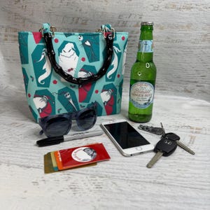 K&ouml;nnte beinhalten: Eine t&uuml;rkisgr&uuml;ne Tragetasche mit einem schwarzen Bambusgriff, verziert mit Figuren aus The Nightmare Before Christmas. Au&szlig;erdem abgebildet sind Sonnenbrillen, ein Telefon, eine Flasche Ginger Beer, Schl&uuml;ssel, ein Marker und Karten.