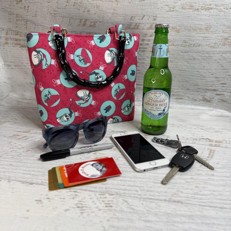 Puede incluir: Un bolso tote rojo con un asa de bamb&uacute; negro, gafas de sol, un tel&eacute;fono, llaves, un rotulador y una botella de cerveza de jengibre. El bolso tiene un estampado de c&iacute;rculos turquesa con ilustraciones sobre fondo rojo.