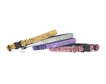 May include: A set of four pet collars featuring a glittery finish in gold, silver, purple, and pink. Each collar includes a black plastic side-release buckle, a metal D-ring for leash attachment, and a black fabric lining on the inner side.
