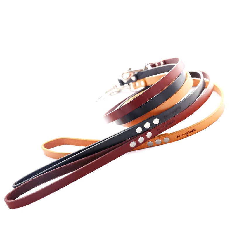 May include: Four leather dog leads in different colours, including brown, tan, black, and burgundy. The leads are stacked on top of each other and have silver metal clips. The leads are engraved with the text "mimigreen".