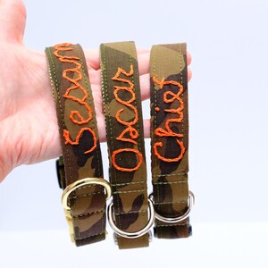May include: Three dog collars with a camouflage design. Each collar is embroidered with a different name in orange thread: "Sesan", "Oscar", and "Chief". The collars have metal fixings and are displayed against a white backdrop.