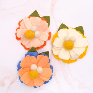 May include: Three felt flower hair clips in different colors. The flowers are layered with a peach, orange, and yellow center. The flowers have white felt balls in the center.