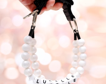 Puede incluir: Un collar personalizado para perros con cuentas blancas y redondas y el nombre "LUCILLE" escrito con cuentas de letras. El collar tiene una correa de nailon negro y herrajes de metal plateado.