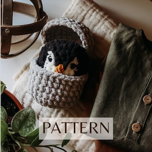 Peut inclure: Un jouet amigurumi en forme de pingouin crocheté dans un panier gris en forme d'œuf. Le jouet a une fourrure noire et blanche et un bec orange. Le panier est sur une surface texturée blanche et beige. Le mot "PATTERN" est imprimé en brun sur un rectangle blanc.