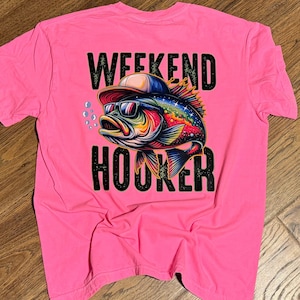 May include: Pink t-shirt featuring a graphic of a fish wearing sunglasses and a hat. The text "Weekend Hooker" is printed below the fish.