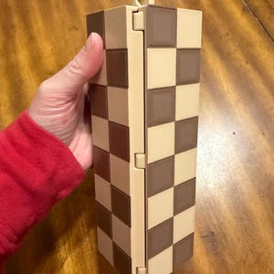 May include: A rectangular, hinged box with a checkerboard pattern in shades of brown and beige. The box is held upright, showcasing its design. The box has a small loop at the top. The box is made of a rigid material.