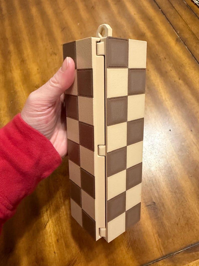 May include: A rectangular, hinged box with a checkerboard pattern in shades of brown and beige. The box is held upright, showcasing its design. The box has a small loop at the top. The box is made of a rigid material.