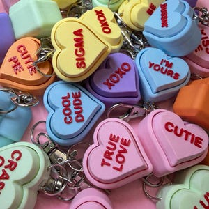 May include: A collection of colorful heart-shaped keychains in various pastel shades, including yellow, blue, pink, and green. Each keychain features a different phrase in red, such as "True Love," "Cool Dude," and "XOXO."