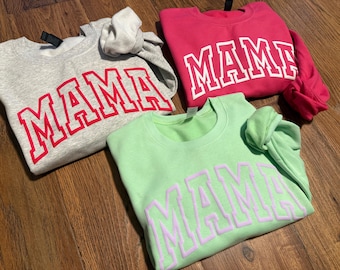 May include: Three crewneck sweatshirts in gray, pink, and light green. Each sweatshirt has the word "MAMA" printed across the front in a bold, collegiate-style font. The gray sweatshirt has red lettering, the pink has white, and the green has lavender.