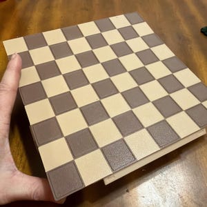May include: A brown and tan chess board with a checkered pattern. The board is rectangular and has a border. The squares alternate between light tan and brown. The board is on a wooden surface.