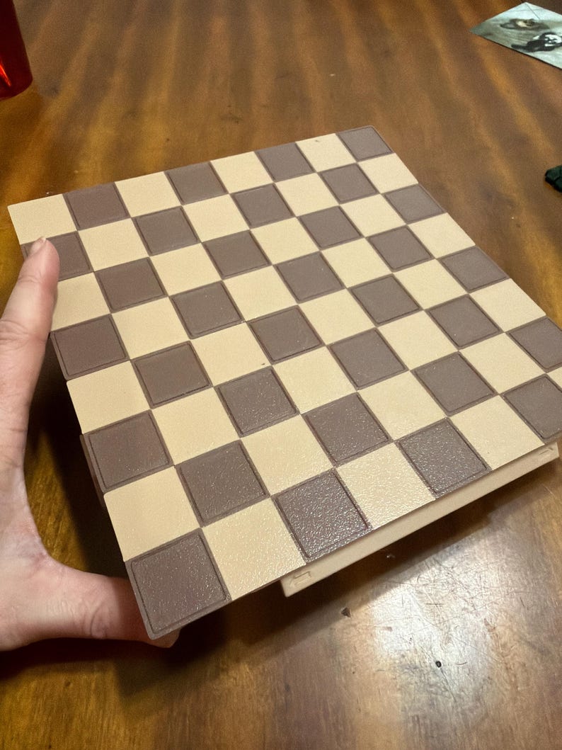 May include: A brown and tan chess board with a checkered pattern. The board is rectangular and has a border. The squares alternate between light tan and brown. The board is on a wooden surface.
