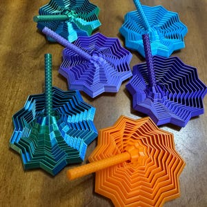 May include: A collection of colorful, star-shaped 3D-printed tops in shades of blue, green, purple, and orange. Each top has a central, textured handle. The tops are arranged on a wooden surface.