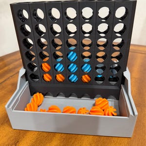May include: A 3D-printed Connect Four game. The game board is black with circular openings. The game pieces are orange and blue, and are stored in a gray tray. The game is set on a wooden surface.