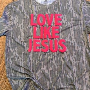 May include: A camouflage-patterned t-shirt with the words "LOVE LIKE JESUS" in bright pink lettering. The shirt has a crew neck and short sleeves. The fabric has a mottled pattern of gray, brown, and white.