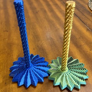 May include: Two decorative candle holders. One is blue, the other is gold. Each has a starburst base and a twisted, textured stem. The bases are layered, creating a dimensional effect. The candle holders are made of plastic.