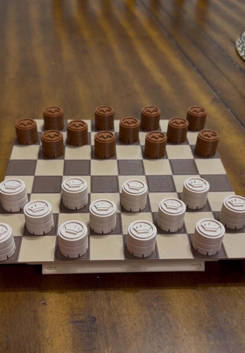 May include: A checkerboard with brown and beige squares. Brown checkers with a circular design are on one side, and beige checkers with a crown design are on the other. The game is set up on a wooden table.