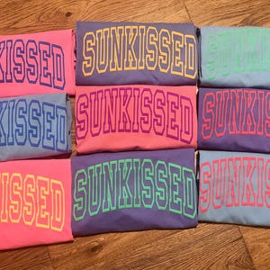 May include: A stack of eight t-shirts with the word "SUNKISSED" printed in different colors on each shirt. The shirts are arranged in a rainbow pattern, with the colors pink, blue, purple, green, and light blue.