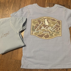 May include: Two light blue t-shirts with the text "Local Boy" printed on them. One shirt has a camouflage design with a dog silhouette.