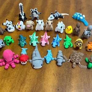 May include: A collection of colorful, handcrafted animal figurines, including a skunk, raccoon, panda, octopus, and turtle. The figurines are arranged on a wooden surface, showcasing a variety of shapes and sizes.