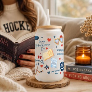 May include: A frosted glass tumbler with a wooden lid and straw, decorated with hockey-themed graphics and text. The tumbler sits on a wooden coaster next to a lit candle and books. The text on the tumbler reads "In my Hockey Romance Era."