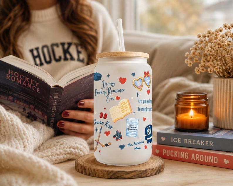 May include: A frosted glass tumbler with a wooden lid and straw, decorated with hockey-themed graphics and text. The tumbler sits on a wooden coaster next to a lit candle and books. The text on the tumbler reads "In my Hockey Romance Era."