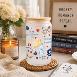 May include: A frosted glass tumbler with a bamboo lid and straw, decorated with hockey-themed graphics and text. The design includes hearts, books, hockey sticks, and the phrase "In my Hockey Romance Era." A candle and books are in the background.