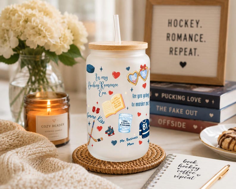 May include: A frosted glass tumbler with a bamboo lid and straw, decorated with hockey-themed graphics and text. The design includes hearts, books, hockey sticks, and the phrase "In my Hockey Romance Era." A candle and books are in the background.