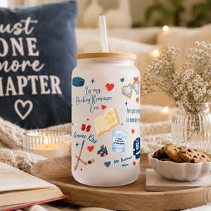 May include: A white glass tumbler with a bamboo lid and a straw, decorated with hockey-themed illustrations and text. The tumbler has the words "In my Hockey Romance Era" and other phrases. A navy blue pillow with the text "just ONE more CHAPTER" is in the background.