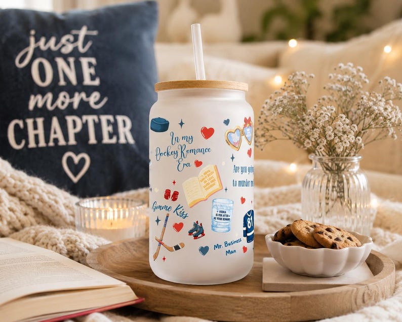 May include: A white glass tumbler with a bamboo lid and a straw, decorated with hockey-themed illustrations and text. The tumbler has the words "In my Hockey Romance Era" and other phrases. A navy blue pillow with the text "just ONE more CHAPTER" is in the background.