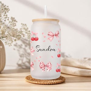 Könnte beinhalten: Ein mattierter Glasbecher mit Bambusdeckel und Strohhalm. Der Becher ist mit rosa Schleifen, Kirschen und dem Namen "Sandra" in Schreibschrift verziert. Er steht auf einem runden, gewebten Untersetzer.