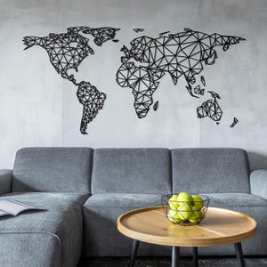 Op de afbeelding: Een zwarte geometrische wereldkaart wanddecoratie, gemonteerd op een lichtgrijze muur boven een grijze hoekbank. Een ronde houten salontafel met een draadmand met groene appels staat voor de bank.