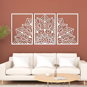 Könnte beinhalten: Dreiteiliges Wandkunst-Set mit weißem Mandala-Design. Jedes Paneel zeigt ein detailliertes, symmetrisches Blumenmuster in einem rechteckigen Rahmen. Das Kunstwerk ist an einer rotbraunen Wand über einem weißen Sofa und einem runden Holztisch angebracht.
