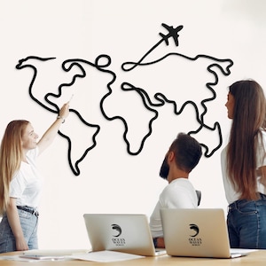 May include: A black metal world map wall art with a stylized airplane design. The map is mounted on a white wall. Three people are in the foreground, with two laptops visible on a wooden table.