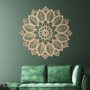 Op de afbeelding: Een groot, ingewikkeld gesneden houten mandala-wandkunstwerk. De mandala heeft een symmetrisch bloemenontwerp met blad- en hartdetails. Het kunstwerk is lichtbruin van kleur en is gemonteerd op een donkergroene muur boven een groene bank.