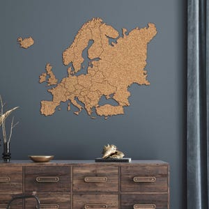 Peut inclure: Une carte en liège texturée de l'Europe, affichant les frontières des pays, fixée sur un mur bleu foncé. En dessous, un meuble en bois avec des tiroirs, un bol doré et des objets décoratifs.