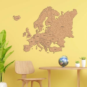 Peut inclure: Une grande carte en liège marron de l'Europe est fixée sur un mur jaune. La carte comprend les frontières et les noms des pays. Un petit globe et des plantes en pot sont sur une table en bois devant la carte. Une chaise en bois est à gauche.